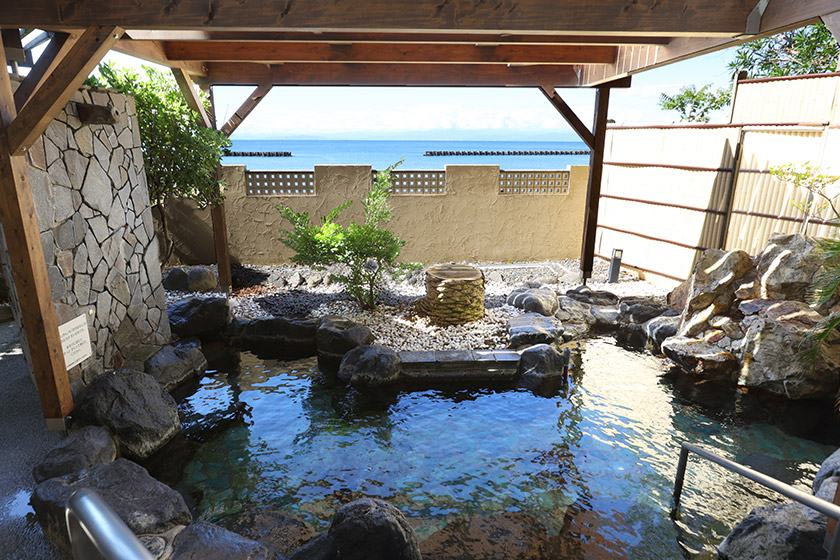 1階 男性露天風呂：野趣のある岩風呂にマリンサイドならではのウッドルーフや白壁、フェニックスの樹の組み合わせがおしゃれです。